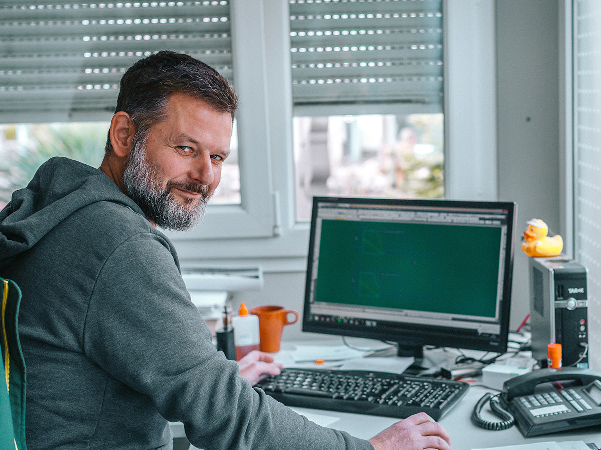 Aufnahme eines Mitarbeiters - BGS GmbH Ein Mitarbeiter sitzt am Schreibtisch und arbeitet, während er in die Kamera lächelt. BGS GmbH